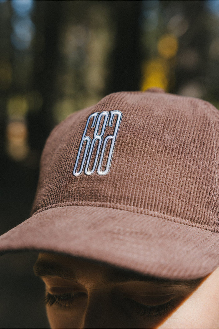A close-up of a person's face wearing the 686 Mid-Pro Ghost Cord Unstructured 5 Panel Hat by 686, featuring brown corduroy fabric and "602" embroidered in white and blue on the front. The polyester cotton woven hat stands out against a blurred background with hints of green and yellow.