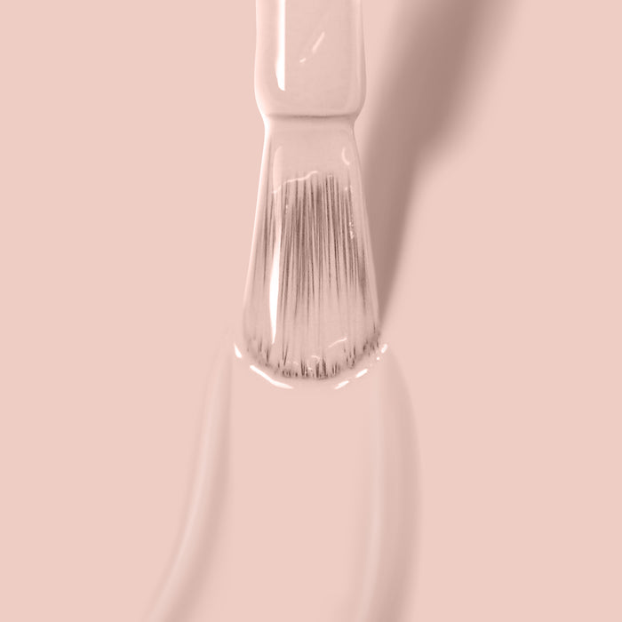 Close-up of a white paintbrush applying LONDONTOWN's Uncovered light pink paint on a smooth surface with a soft shadow in the background, blending seamlessly like nude polish.