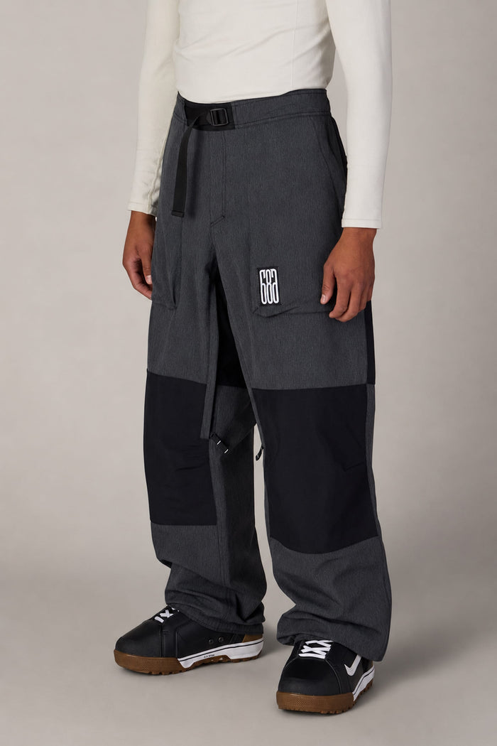 A person is wearing the 686 Ghost™ 2.5L Pant by 686—loose-fitting, color-blocked in gray and black with a signature patch—paired with a white long-sleeve shirt and black athletic shoes, standing against a plain light gray background.