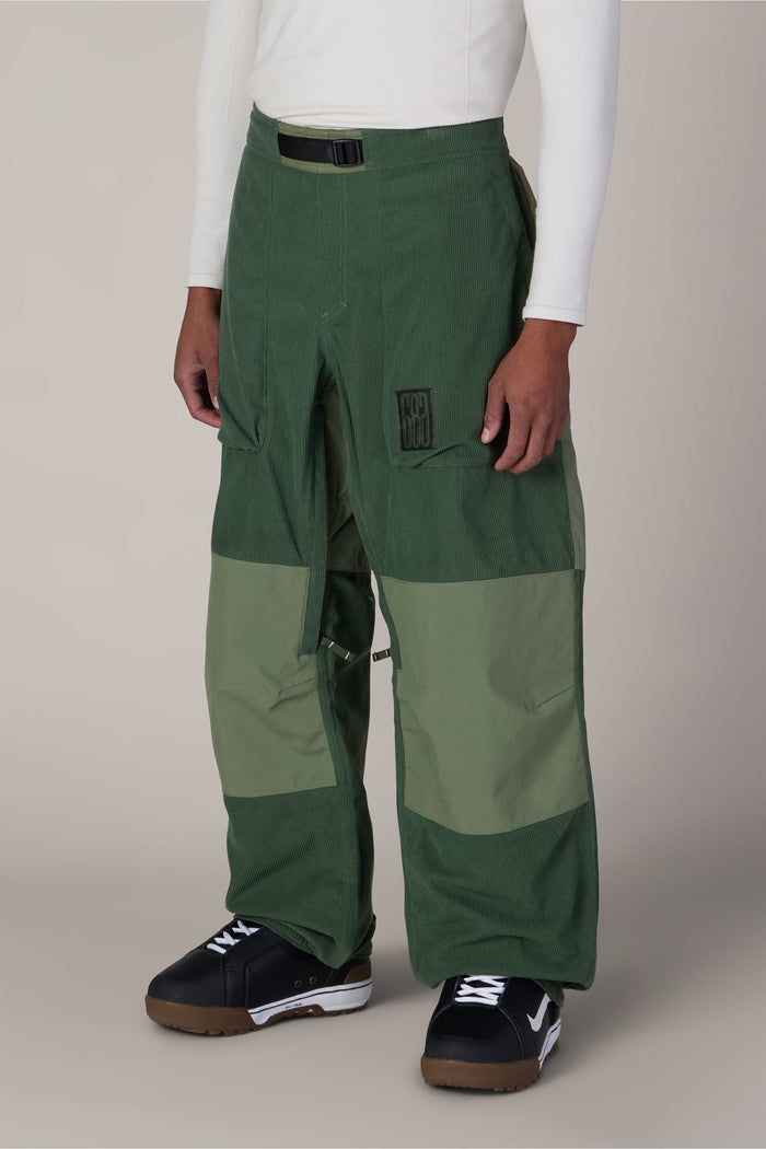A person is wearing the 686 Ghost™ 2.5L Pant by 686—two-tone green pants with large front pockets—paired with a black belt, black sneakers with white soles, and a long-sleeve white shirt. The background is plain light gray.
