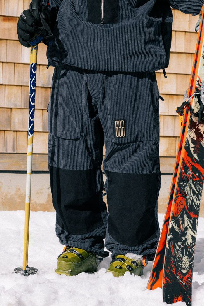 A person stands in the snow, wearing 686 Ghost™ 2.5L Pants and green ski boots, holding a ski pole with a pair of skis resting at their side; only the lower body is visible.