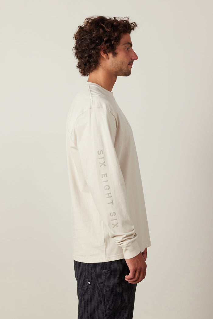 A man with curly hair stands in profile, wearing a 686 High Def Authentic L/S Tee in light beige, made of 100% cotton with ‚ÄúSIX EIGHT SIX‚Äù on the sleeve, paired with dark patterned pants against a plain light background.