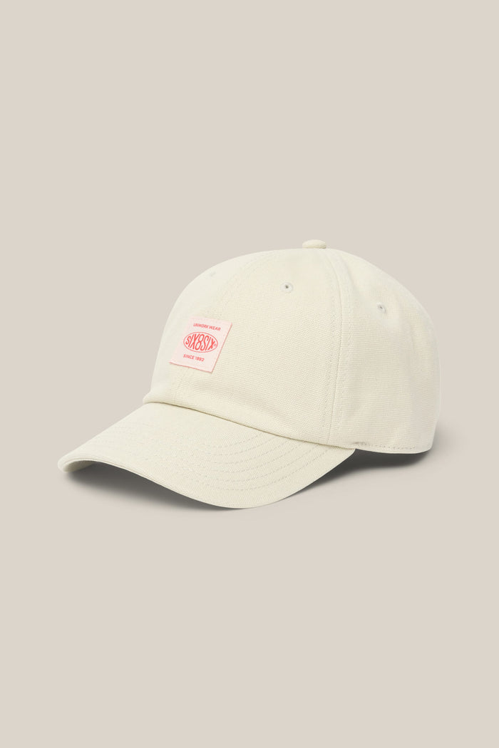Here’s the rewritten sentence using the provided product data: The 686 Low-Pro Unwork Unstructured 6 Panel Hat by 686 features a cream-colored polyester-cotton blend, a curved brim, and a small rectangular pink and white patch on the front, displayed against a plain beige background. This stylish men’s hat combines comfort with understated design.