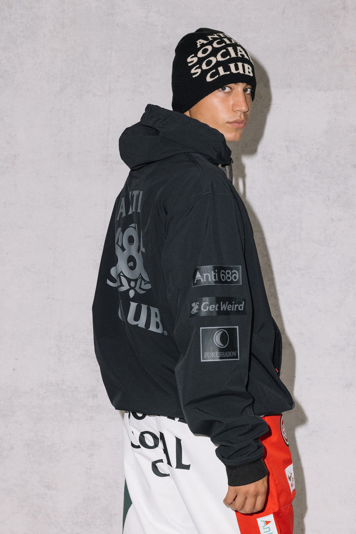 A young man poses sideways in a black 686 Men's ASSC F1 Waterproof Team Hoody, featuring logos and patches, with white and red pants against a plain light-colored wall.