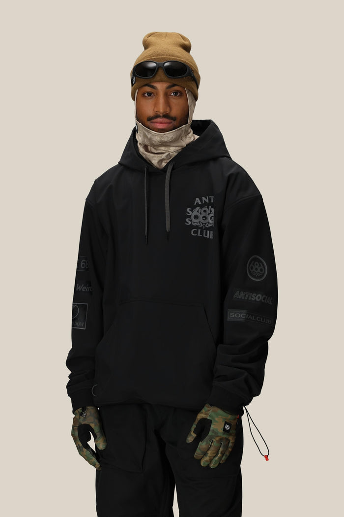 A man wearing a tan beanie with goggles, black pants, camouflage gloves, a beige neck gaiter, and a 686 Men's ASSC F1 Waterproof Team Hoody by 686 stands against a plain light background.