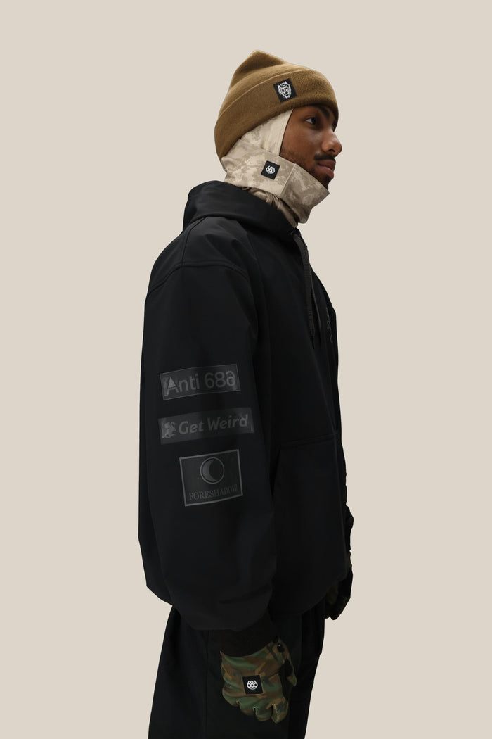 A man wearing the 686 Men's ASSC F1 Waterproof Team Hoody, a tan beanie, beige face covering, and camouflage gloves stands in profile against a plain beige background.