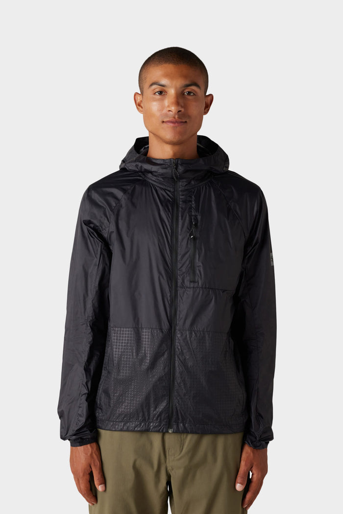 A person stands facing forward against a plain light gray background, wearing the 686 Mens Grid Shell Jacket in black with a zip front and hood, paired with olive-green pants.