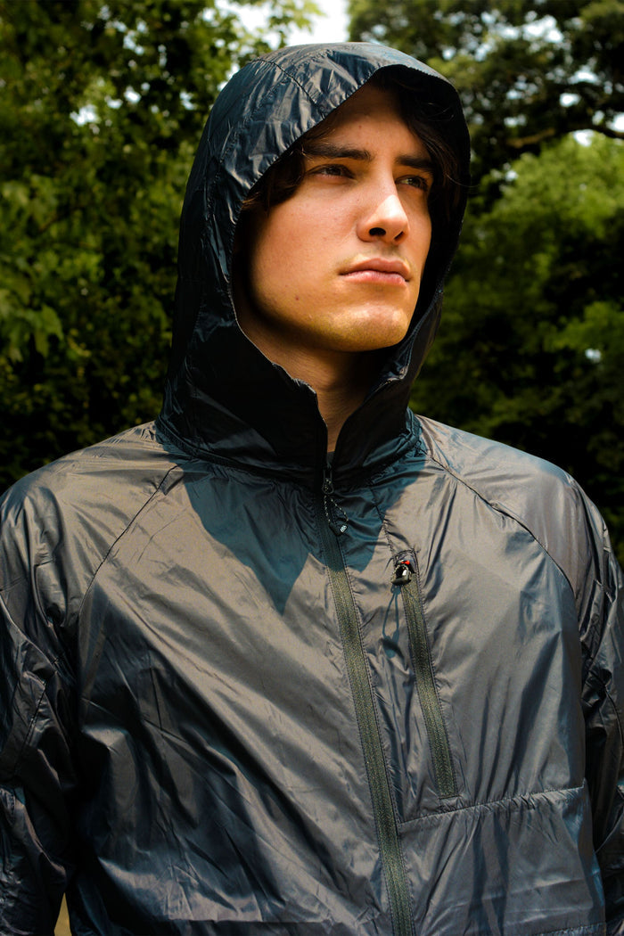 A young man stands outdoors in a 686 Mens Grid Shell Jacket with the hood up, gazing into the distance. Green trees and foliage form a blurred background.