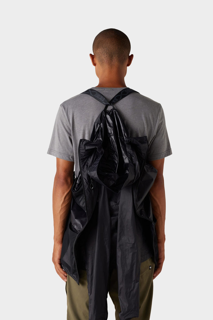 A person is seen from behind wearing olive pants, a grey t-shirt, and the 686 Mens Grid Shell Jacket by 686, a black sleeveless garment draped over their shoulders against a plain light background.