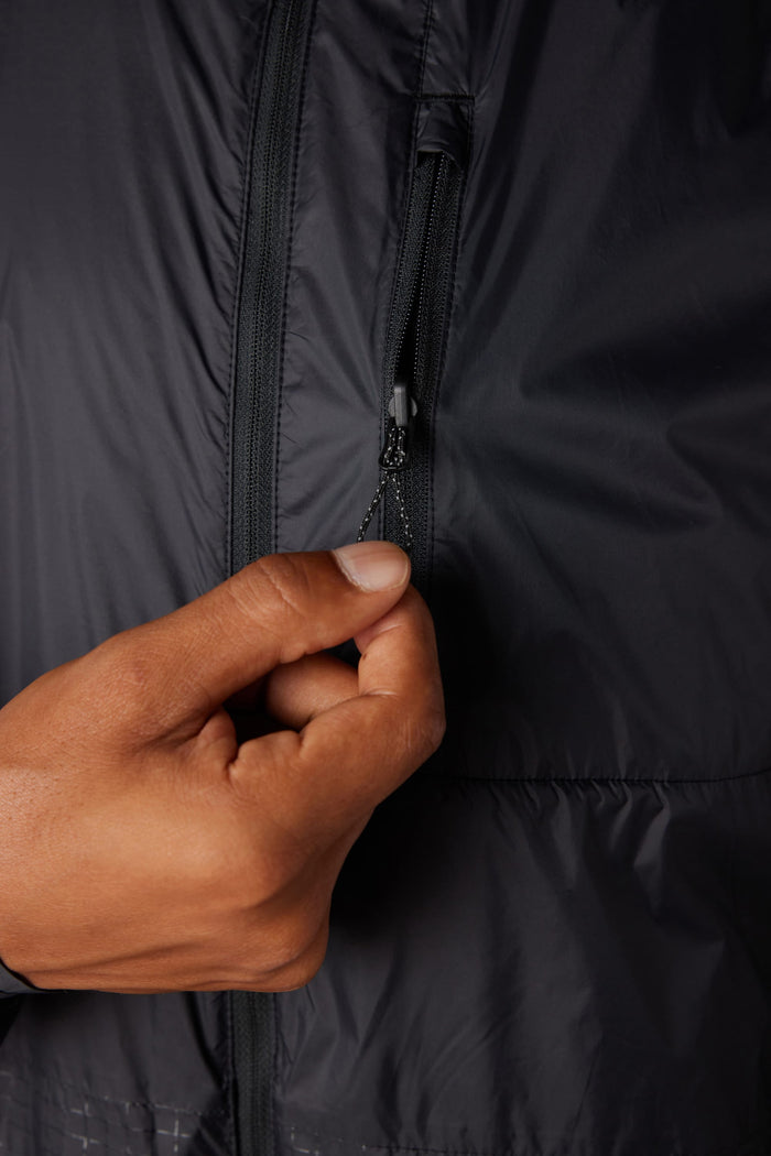 A close-up of a hand pulling the zipper on the small chest pocket of the 686 Mens Grid Shell Jacket. The lightweight, slightly shiny fabric showcases 686‚Äôs signature dark-colored style.
