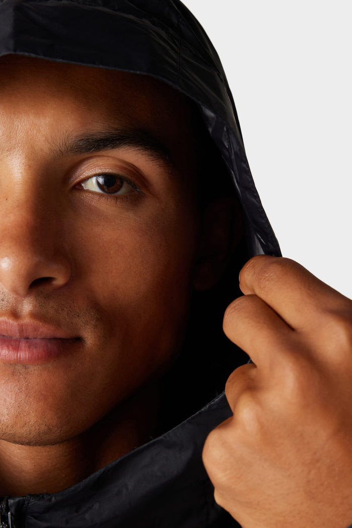Close-up of a person‚Äôs face from the nose up, wearing the 686 Mens Grid Shell Jacket by 686 with the hood up and holding its edge. The plain background is light-colored.
