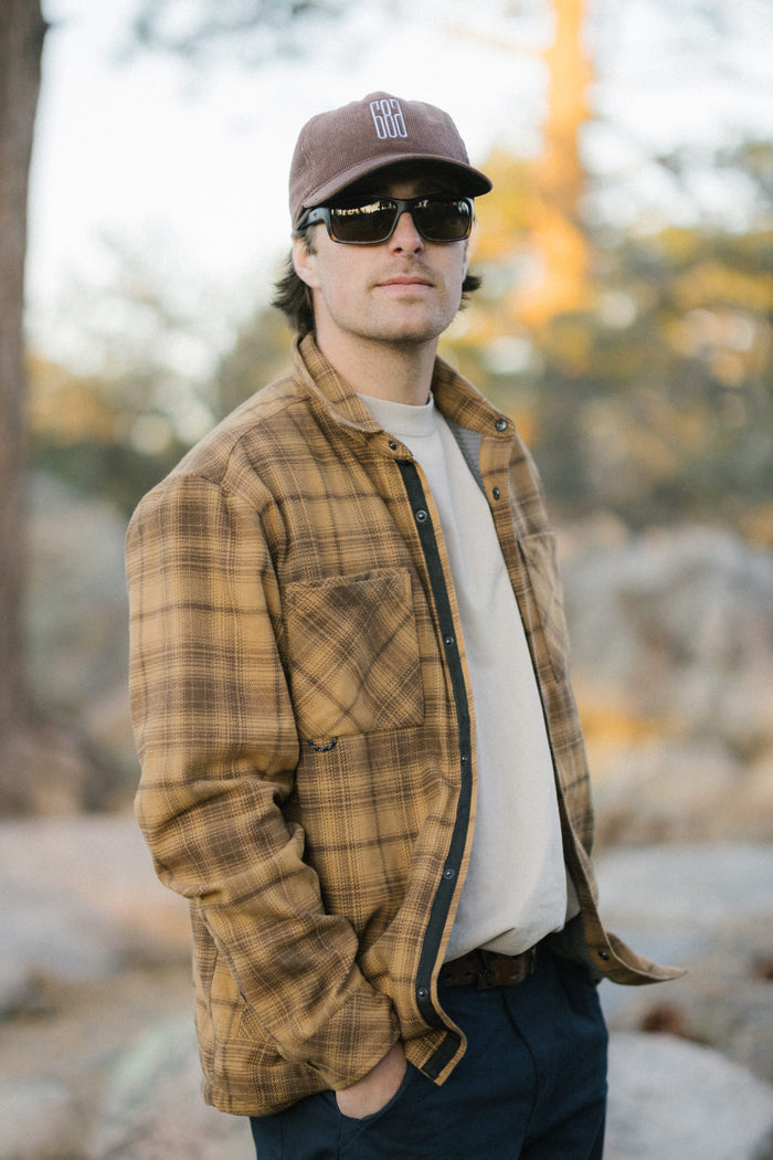 Wearing the 686 Mid-Pro Ghost Cord Unstructured 5 Panel Hat by 686, along with a brown plaid jacket, beige shirt, dark pants, and sunglasses, a person stands outdoors with one hand in their pocket amidst trees and rocks.