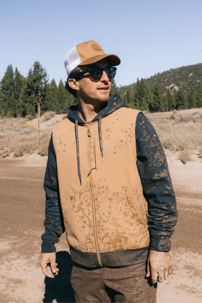 A man wearing sunglasses, the 686 Mid-Pro Wreath Unstructured 5 Panel Mesh Hat by 686 in tan and white, a tan and black hoodie, and brown pants stands outdoors on a muddy trail, with trees and hills in the background. His hoodie and pants are splattered with mud.
