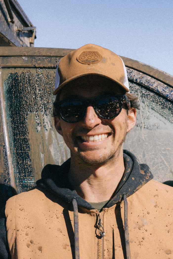 A person wearing the 686 Mid-Pro Wreath Unstructured 5 Panel Mesh Hat by 686, along with sunglasses and a tan vest, smiles in front of a mud-splattered vehicle on a sunny day.
