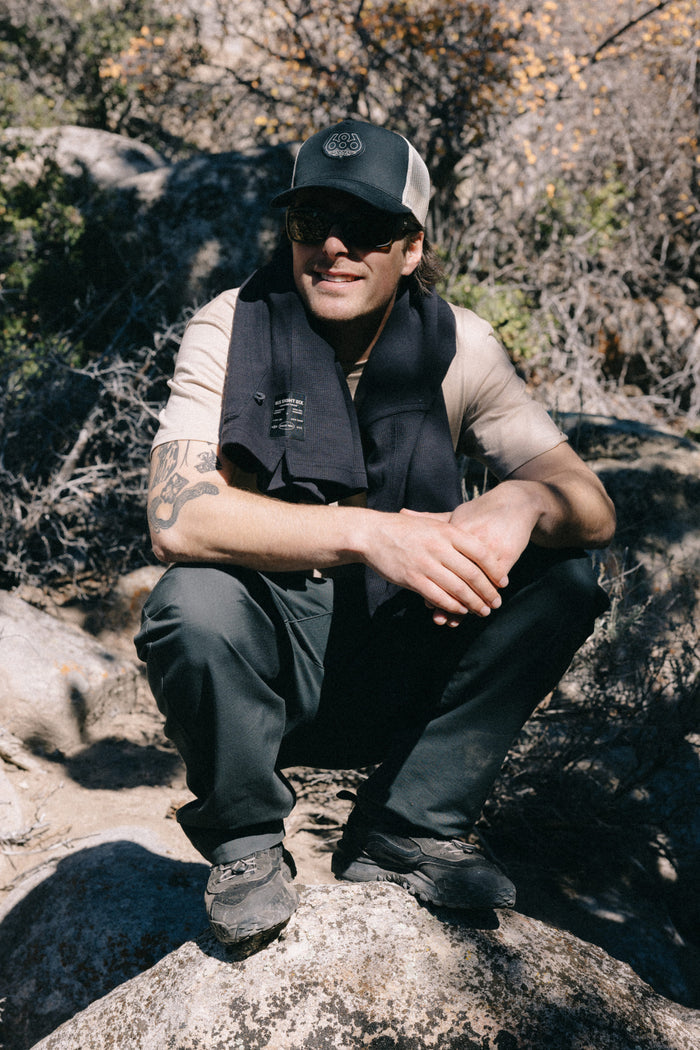 A person wearing the 686 Mid-Pro Wreath Unstructured 5 Panel Mesh Hat by 686, along with sunglasses and outdoor attire, squats on a rock in a sunlit, rocky landscape with sparse vegetation.