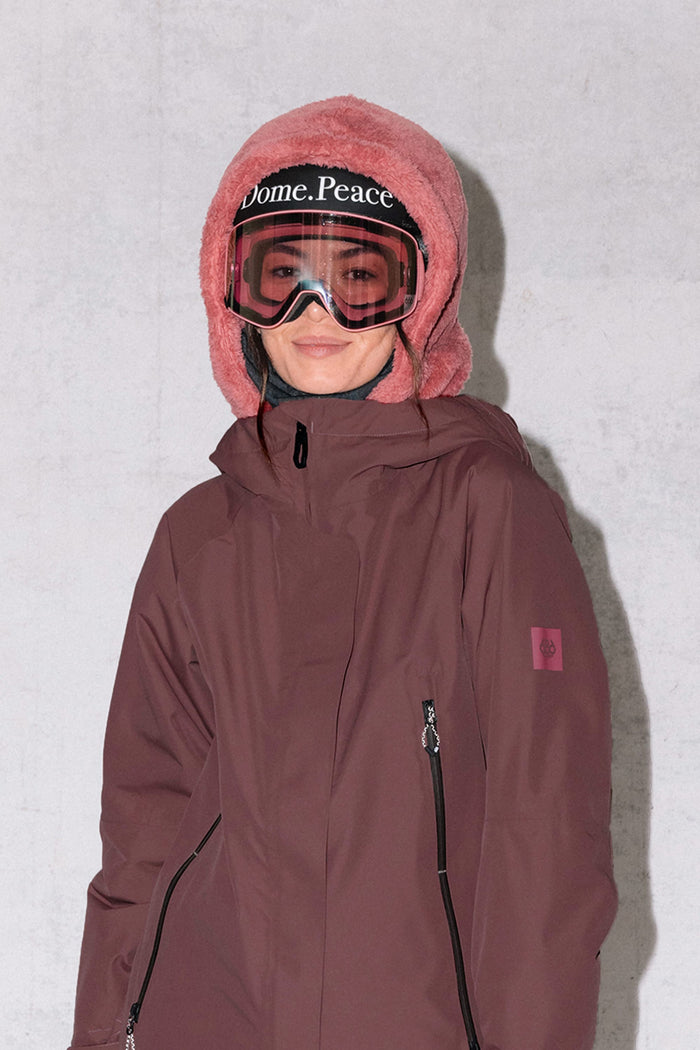 A person in a maroon winter jacket and pink fuzzy hat stands by a light gray wall, smiling, with the 686 Tarmac Sherpa Hood by 686‚Äîa polyester knit hood‚Äîvisible under the hat, and wearing ski goggles labeled Dome. Peace.