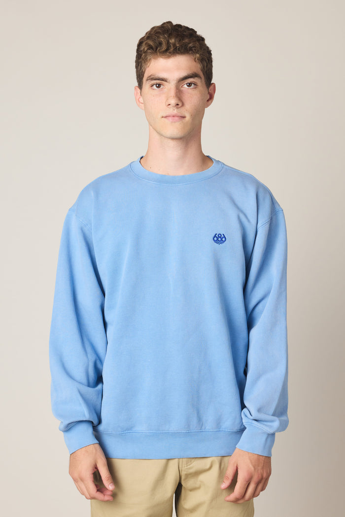 A young man with short curly hair wears the 686 Wreath Relaxed LS Crewneck‚Äîa light blue 100% cotton sweatshirt with a small embroidered logo on the chest‚Äîpaired with beige pants, standing against a plain light background.
