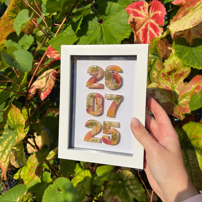 Framed artwork with colorful numbers held by a hand among green leaves