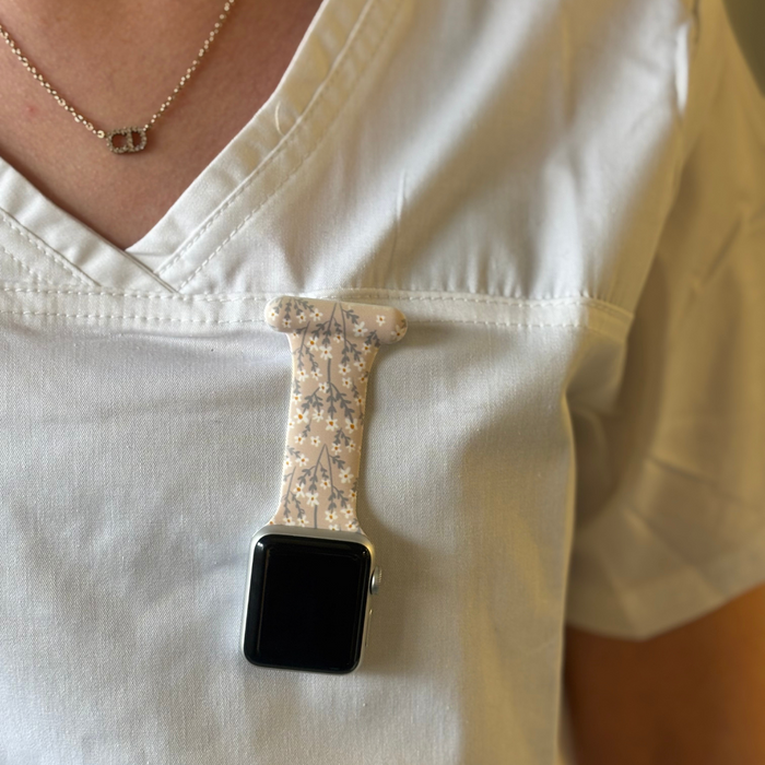 Coconut Lane Apple Watch fob strap in nude floral design clipped upside down on a light-colored scrub top pocket.