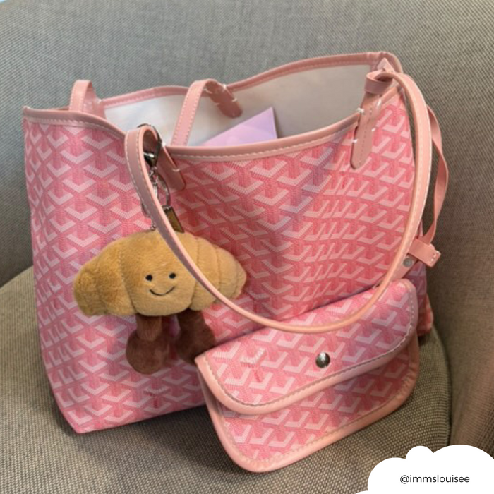Pink patterned tote with a croissant-shaped bag charm and matching pouch, resting on a grey fabric chair.