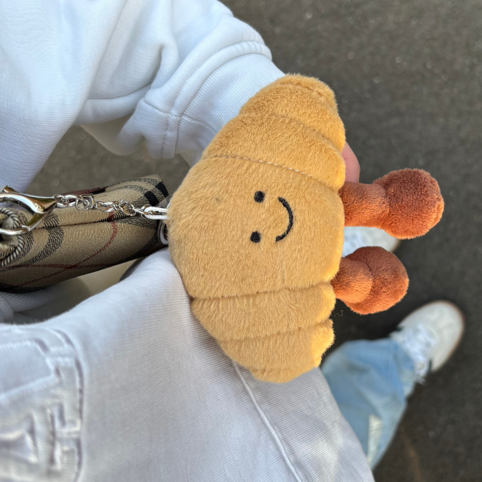 A person in a white jacket and blue jeans holds a smiling croissant-shaped bag charm from Coconut Lane.