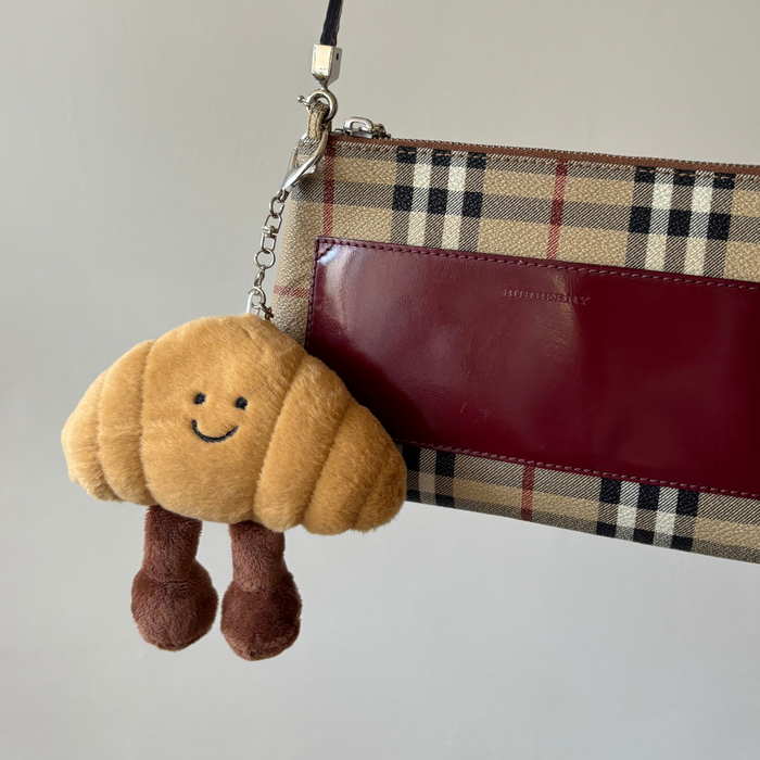 Smiley croissant bag charm with plaid and burgundy details, attached to a Parisian-inspired accessory.