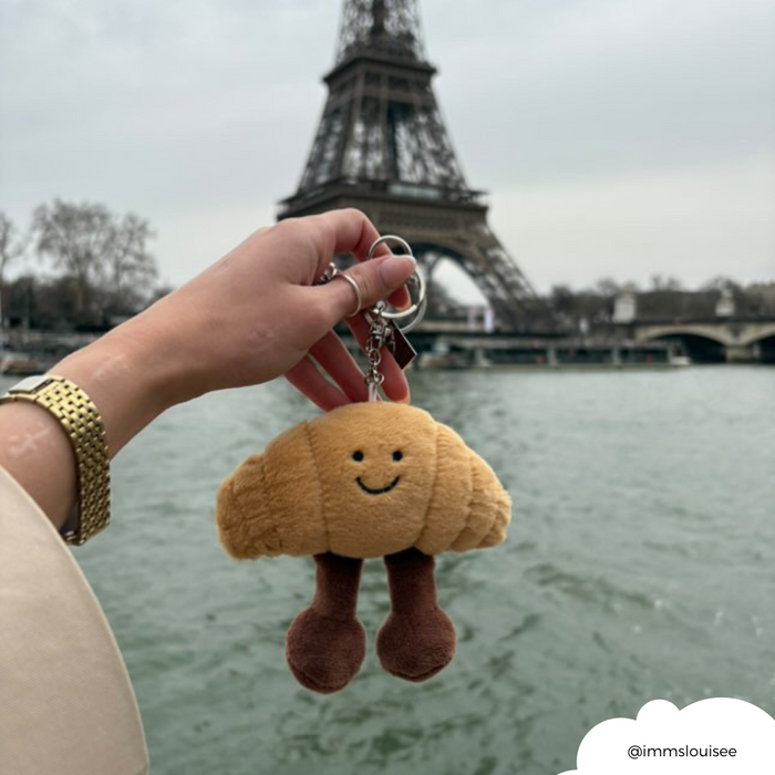 Hand holding a croissant-shaped bag charm with gold chain in front of the Eiffel Tower and Seine River.