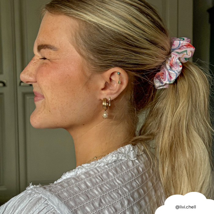 Smiling woman with closed eyes wears a white Dolce Vita satin scrunchie and gold hoop earrings in her blonde ponytail.