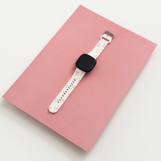 Pink Coconut Lane Fitbit strap with dainty bow pattern and white adjustable band on a matching pink notebook.