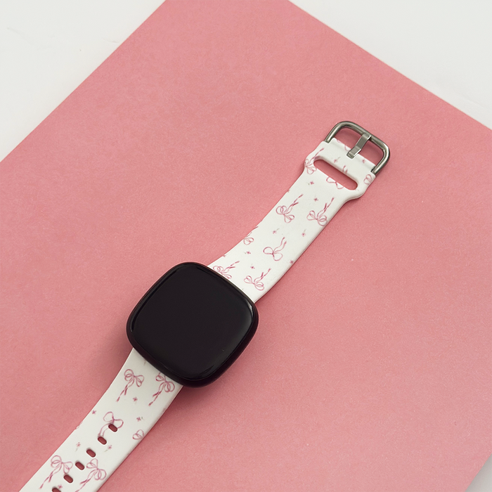 Adjustable Fitbit strap with a white and pink dainty bow pattern, displayed on a pink background.