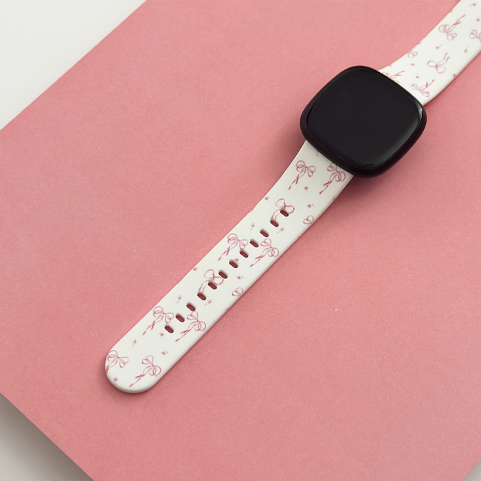 Coconut Lane Fitbit strap in white with pink bow pattern, displayed on a pink surface.