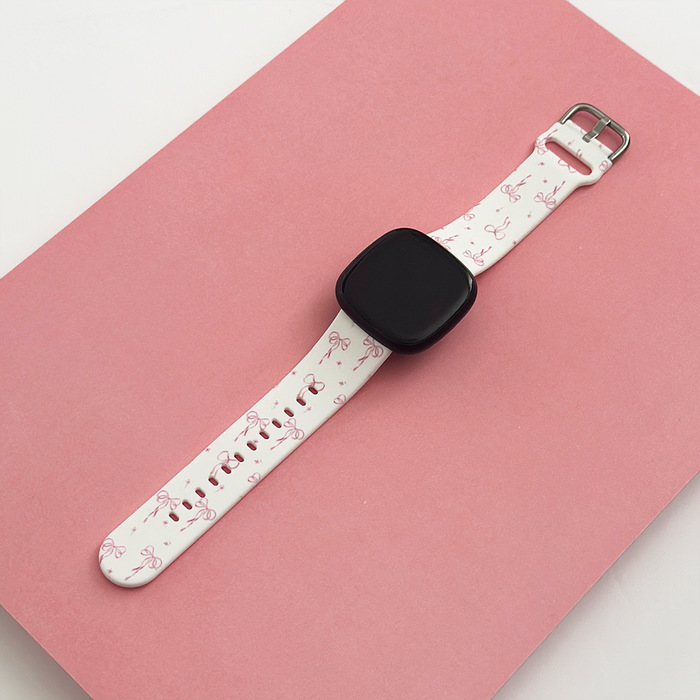 White Fitbit strap with pink bows and ribbons pattern, displayed on a pink and white background.