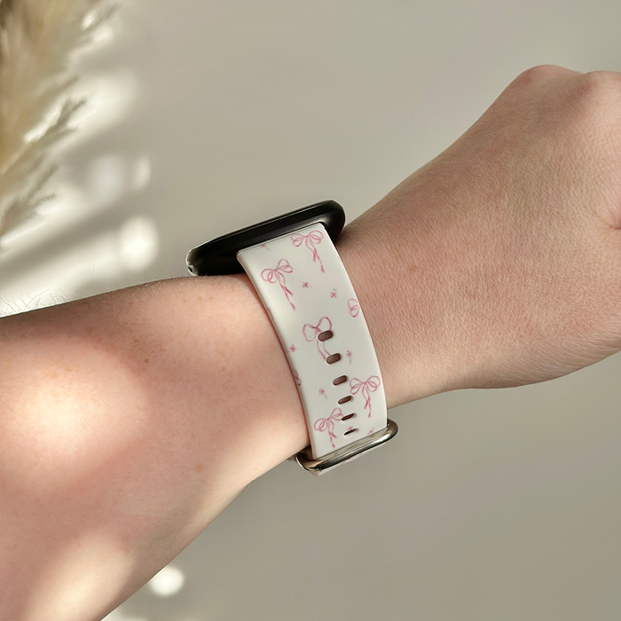 White adjustable Fitbit strap with a pink bow pattern from Coconut Lane, shown worn on the left wrist.