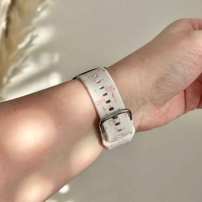 White Coconut Lane Fitbit strap with pink script bows pattern, silver buckle, shown on a wrist.