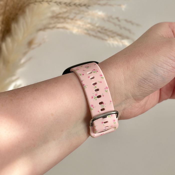 Hand wearing a pink Coconut Lane Fitbit strap with ditsy floral pattern, shown against dried grasses and a neutral backdrop.