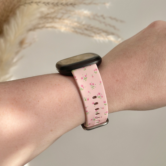 Pink ditsy floral Coconut Lane Fitbit strap on a wrist, with dried grass and a soft neutral backdrop.