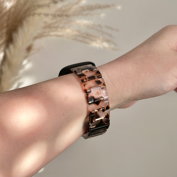 Wrist with Coconut Lane Fitbit Strap in ivory tort pattern, shown close up against a soft beige blurred background.