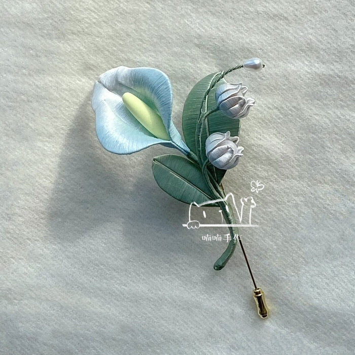 Handmade Chanhua brooch with lifelike light blue calla lily and white lily of the valley displayed on soft off-white fabric