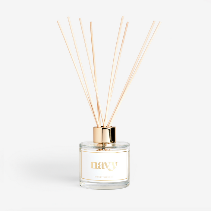 A clear glass reed diffuser with navy branding, containing oil and several light brown reeds, designed to disperse a fragrance.