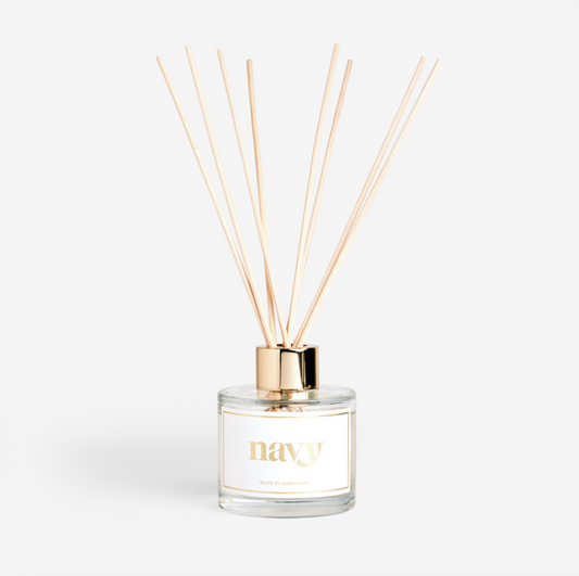 A clear glass reed diffuser with navy branding, containing oil and several light brown reeds, designed to disperse a fragrance.