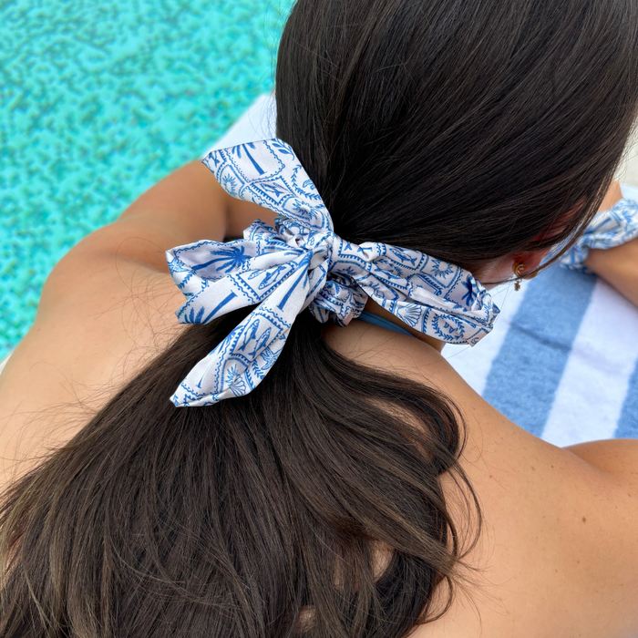 Woman with long dark hair wears a satin bow scrunchie in Malibu Drive pattern, lounging by the pool on a striped towel.