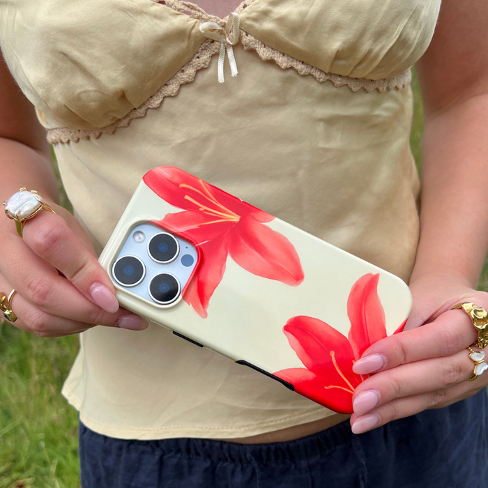 Hand holding phone with Coconut Lane’s Island Girl Ibiza Lily tough case, gold rings, and beige bow top visible.
