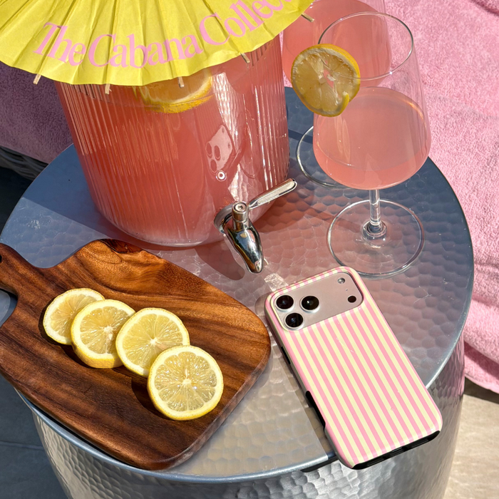 Pink lemonade with a wooden cutting board, sliced lemons, and a tough phone case on a glass table.